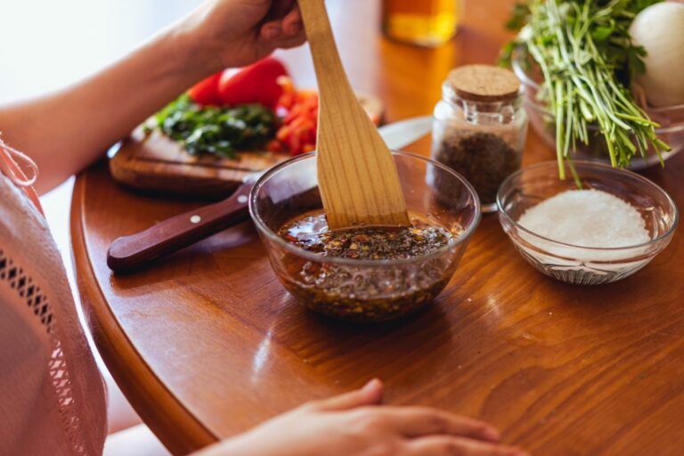 Pessoa preparando uma marinada caseira com ervas, temperos e óleo em uma tigela de vidro, com ingredientes frescos ao fundo
