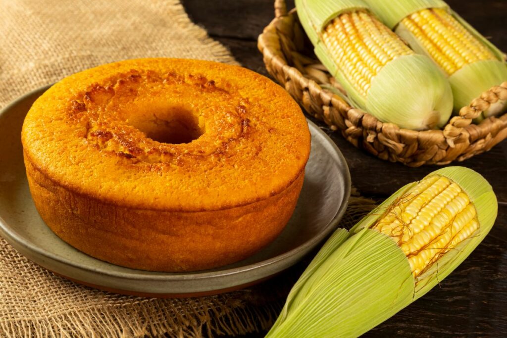 Bolo de fubá dourado sobre prato rústico, cercado por espigas de milho frescas, representando a simplicidade e o sabor das receitas afetivas da culinária atemporal.